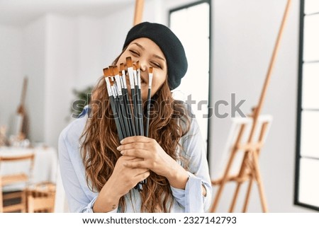 Young beautiful hispanic woman artist covering mouth with paintbrushes at art studio Royalty-Free Stock Photo #2327142793