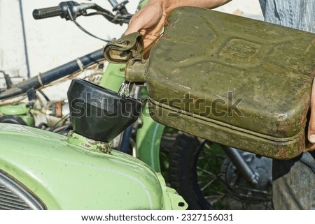 manual refueling with old iron rusty green color large canister of gasoline motorcycle green tank retro motorbike using a large black plastic watering can outdoors at daytime