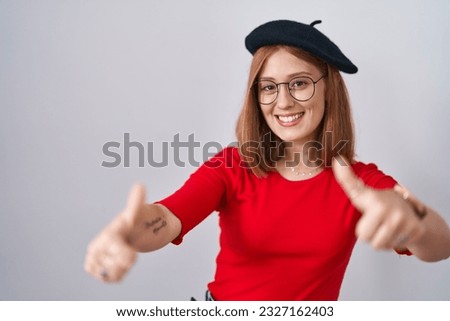 Young redhead woman standing wearing glasses and beret approving doing positive gesture with hand, thumbs up smiling and happy for success. winner gesture. 