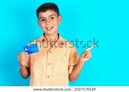 Smiling Little hispanic kid boy wearing orange shirt showing debit card pointing finger empty space
