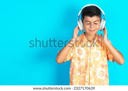 Pleased Little hispanic kid boy wearing orange hawaiian shirt enjoys listening pleasant melody keeps hands on stereo headphones closes eyes. Spending free time with music