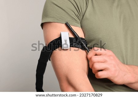 Soldier in military uniform applying medical tourniquet on arm against light grey background, closeup