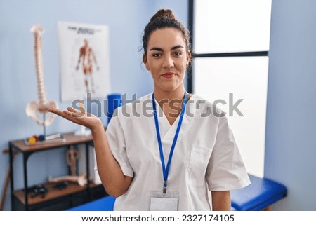 Young hispanic woman working at rehabilitation clinic smiling cheerful presenting and pointing with palm of hand looking at the camera. 