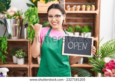 Hispanic young woman working at florist holding open sign pointing thumb up to the side smiling happy with open mouth 