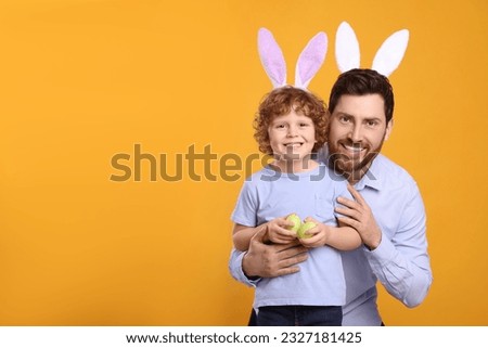 Happy father and son wearing cute bunny ears headbands on orange background. Boy holding Easter eggs, space for text