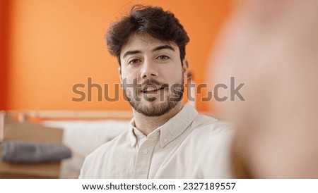 Young hispanic man standing with relaxed expression make selfie picture by smartphone at new home