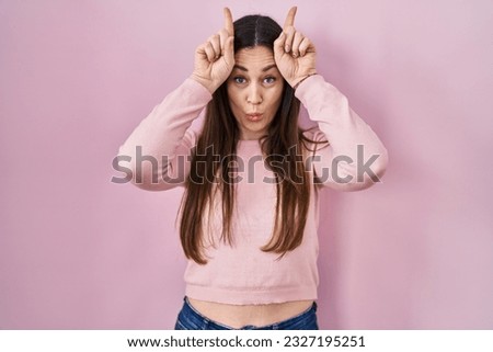 Young brunette woman standing over pink background doing funny gesture with finger over head as bull horns 
