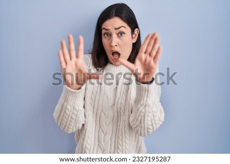 Young brunette woman standing over blue background afraid and terrified with fear expression stop gesture with hands, shouting in shock. panic concept.  Royalty-Free Stock Photo #2327195287