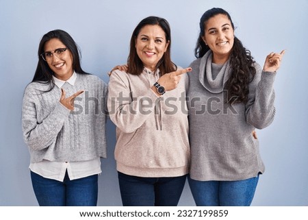 Mother and two daughters standing over blue background with a big smile on face, pointing with hand finger to the side looking at the camera. 
