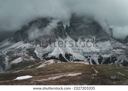 Hiking path with two people walking along a trail in front of a massive mountain with a hut in front in the Dolomites, Italian alps with a storm approaching Royalty-Free Stock Photo #2327201035