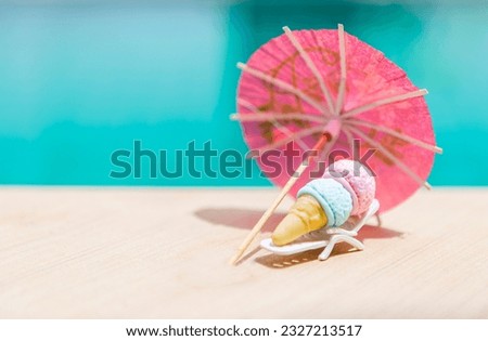 Mini ice cream cone on beach chair with pink paper umbrella on swimming pool edge with space on blurred blue water background, summer outdoor day light, it's summer time for a treat Royalty-Free Stock Photo #2327213517