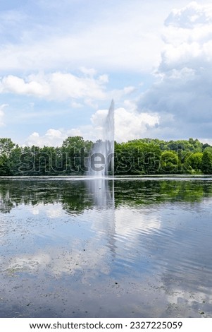 Waterjet and breeze from a fountain on the Echternach lake, hills and green leafy trees on a misty background, reflection on water surface, sunny spring day with abundant clouds in Luxembourg