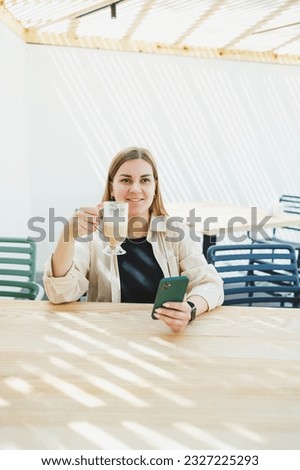 Happy woman sitting at outdoor cafe table and talking on phone with cup of coffee, smiling woman enjoying telecommuting in cafe or studying online