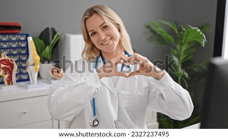 Young blonde woman doctor smiling doing heart gesture with hands at clinic