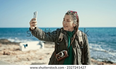 Mature hispanic woman with grey hair tourist wearing backpack smiling taking a selfie picture at seaside