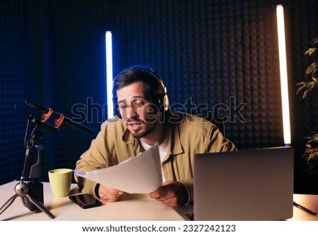 Gesturing radio host with headphones reading news from paper into studio microphone at radio station with neon lights