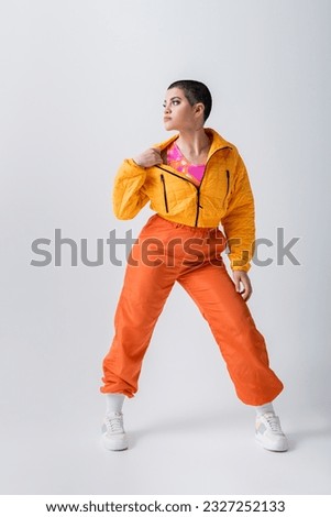 outerwear, casual attire, tattooed and young woman with short hair pulling yellow puffer jacket on grey background, urban fashion, vibrant youth, trendy outfit, stylish look, studio photography Royalty-Free Stock Photo #2327252133