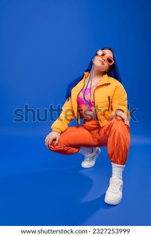 fashion statement, full length of pretty female model with blue hair and trendy sunglasses sitting on haunches on blue background, rebel style, modern fashion, trendy accessory, gen z Royalty-Free Stock Photo #2327253999