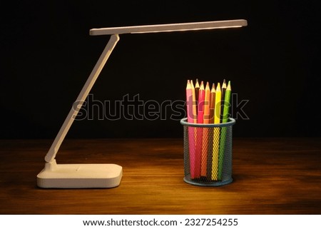 Colored pencils in a stand on a wooden table in the light of a white table lamp.