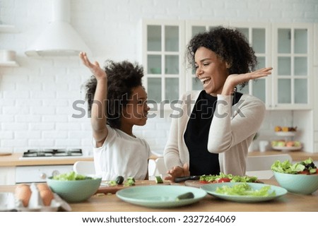 Excited African mom give high five gesture to little daughter family cook together dinner healthy food vegetarian salad. Teach kid, happy motherhood, share cookery skills concept Royalty-Free Stock Photo #2327264069