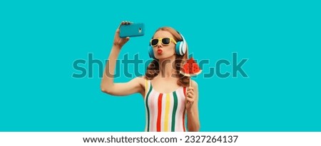 Summer portrait of cheerful happy young woman taking selfie with smartphone in headphones listening to music with juicy lollipop or ice cream shaped slice of watermelon on blue background