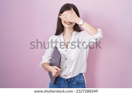 Young caucasian woman holding laptop smiling and laughing with hand on face covering eyes for surprise. blind concept. 