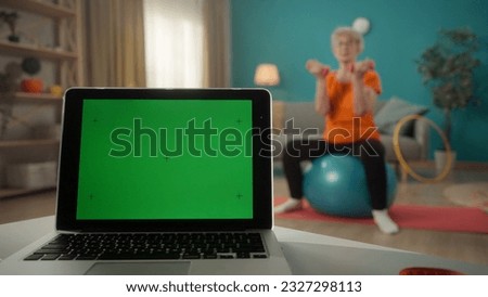 A laptop with a green screen on a table on a blurred background in the living room close up. An elderly woman is sitting on a fitness ball with dumbbells. Advertising area, workspace mock up.