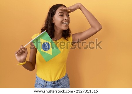 Young hispanic woman holding brazil flag very happy and smiling looking far away with hand over head. searching concept. 