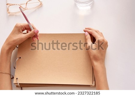 Woman is writing by her left hand on a craft paper paper. Mockup album. Glasess and glass of water. Conceptual photo of international left handers day. Copy space. Top view. Royalty-Free Stock Photo #2327322185