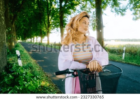 Portrait of young caucasian woman with blond hair riding on bicycle among agricultural fields in countryside in summer day.