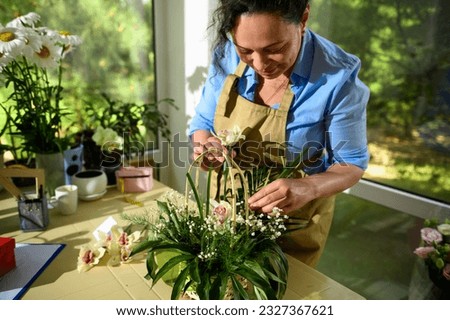 Florist inserts an orchid flower into a soaked foam inside a wicker basket while arranging bouquet, with palm leaves and gypsophila flowers in floral design studio. Creative people and small business