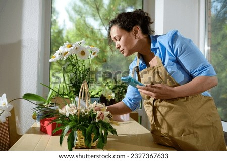 Beautiful inspired multi-ethnic female florist, flower designer entrepreneur in beige apron holding mobile phone, working with flower arrangements for special festive events, in a floral design studio