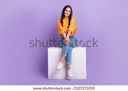 Full length photo of gorgeous friendly cute girl dressed yellow shirt denim pants sit on white cube isolated on purple color background