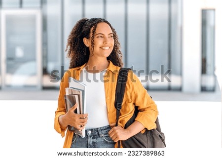 Happy female student. Positive female student, brazilian or hispanic nationality, with a backpack, holding books and notebooks in her hand, stand near the university campus, looks away and smiling Royalty-Free Stock Photo #2327389863