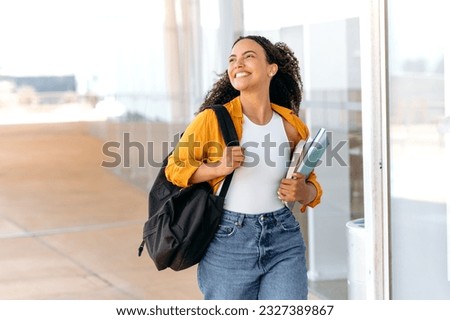 Happy lovely curly haired brazilian or hispanic female student, with a backpack, hold books and notebooks in her hand, walking near the university campus, looks away and smile, finished school day