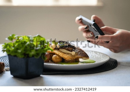 Roasted branzino with marinated mushrooms and butter potatoes on the plate. A woman taking photo with smart phone of the food.  Posting and sharing food pictures on social media.