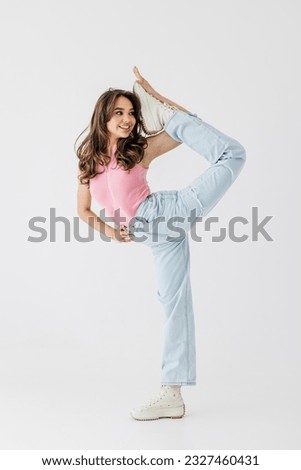 Full length photo of an attractive lady with her leg raised, standing in a split, flexible body, imitating a conversation on the phone, wearing casual jeans and a shirt, on a white background
