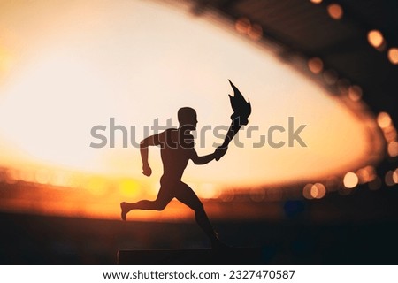 Silhouette of Male Athlete Carrying the Torch Relay, Illuminating the Modern Track and Field Stadium. A Captivating Snapshot for the Summer Game 2024 in Paris. Royalty-Free Stock Photo #2327470587