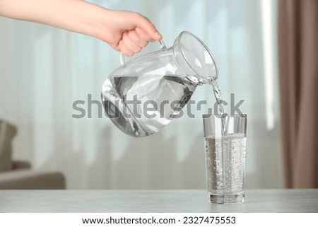 Woman pouring water from jug into glass at white table indoors, closeup Royalty-Free Stock Photo #2327475553