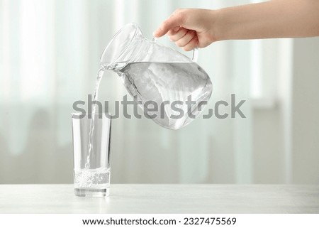 Woman pouring water from jug into glass at white table indoors, closeup Royalty-Free Stock Photo #2327475569
