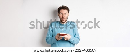 Excited young man making wish on birthday cake with lit candle, celebrating bday, standing on white background.