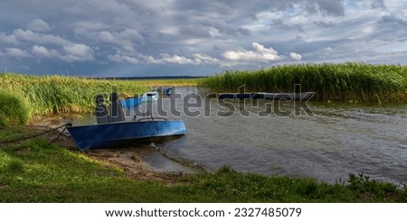 fishing boats parked in a small lake bay overgrown with reeds under a dramatic cloudy sky in windy weather before rain