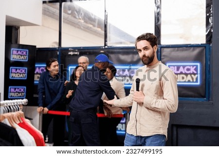 Journalist holding mic gesturing while doing Black Friday reportage in fashion boutique. TV reporter talking over microphone while standing against crowd of shoppers eager to start shopping Royalty-Free Stock Photo #2327499195