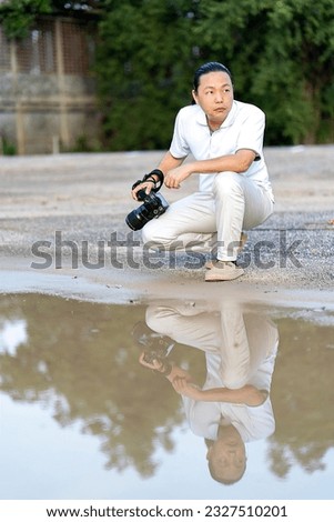 Asian man holds a big black mirrorless camera medium format type on cement asphalt ground in front of water reflex.
