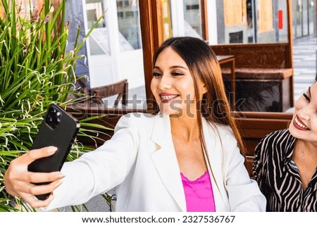 Two women smiling while taking selfies with mobile phone at a cafe. Technology and friendship concept.