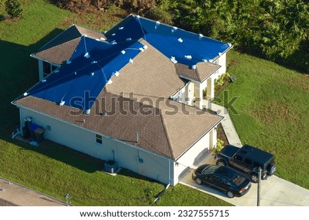 Aerial view of damaged in hurricane Ian house roof covered with blue protective tarp against rain water leaking until replacement of asphalt shingles Royalty-Free Stock Photo #2327555715