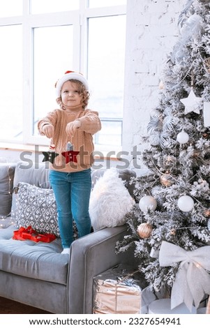 cute happy little girl with curls in Santa hat hangs toys and decorate with ornaments Christmas tree at home, New Year and Christmas family concept