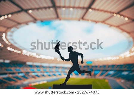 Silhouette of male athlete holding torch relay. Modern Track and Field Stadium in background. Photo for Summer game in Paris