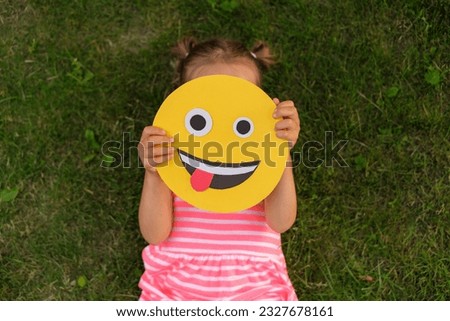 A close-up of crazy smile face making faces and showing his tongue in the hands of a small child lying on the grass of a lawn in a park