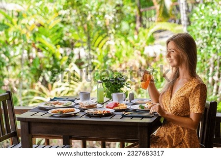 Happy young woman sitting at table, savoring delicious breakfast by the jungle. Concept of luxury resort and vacation. Royalty-Free Stock Photo #2327683317